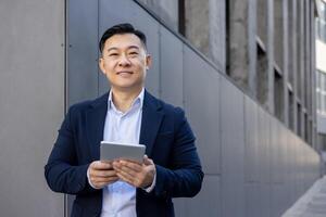 professionell asiatisk manlig verkställande bläddring på en digital läsplatta medan stående utomhus med en trevlig uttryck. foto