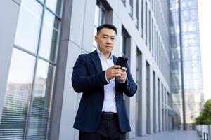 allvarlig asiatisk affärsman stående på de gata nära de kontor med en mobil telefon, skriver en meddelande, framställning en ringa upp, väntar för ett utnämning. foto