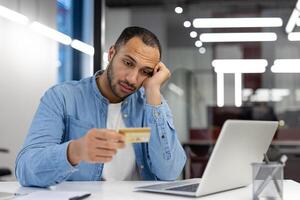 en man visas orolig som han innehar en kreditera kort och stirrar på hans bärbar dator skärm, eventuellt som handlar om med finansiell frågor. foto