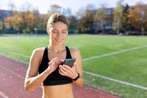 ung sportig kvinna idrottare och löpare stående leende i stadion i sportkläder och använder sig av telefon. foto