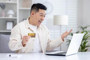 Lycklig ung asiatisk man innehav kreditera kort i hand och känslomässigt Lycklig ser på bärbar dator skärm. foto
