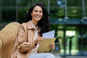 leende ung kvinna med lockigt hår Sammanträde utanför ett kontor, tar en ha sönder med dokument i hand. foto
