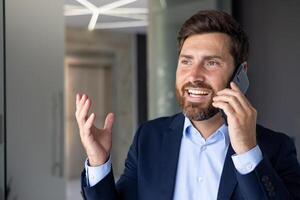 närbild Foto av framgångsrik mogna affärsman talande på de telefon, man i företag kostym leende belåtet på arbetsplats inuti kontor, hört klienter avlägset.