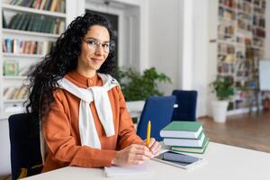 porträtt av ung skön latin amerikan lärare, kvinna i glasögon leende och ser på kamera, undervisning studenter och elever Sammanträde i klassrum bland böcker. foto