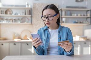 en ung asiatisk kvinna försöker till do en bank drift, användningar en telefon och en Bank kreditera kort, besviken och olycklig med de resultat foto