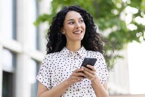 en ung skön latin amerikan kvinna med lockigt hår promenader genom de stad med en telefon i henne händer, en kvinna användningar ett Ansökan på en smartphone, bläddrar internet sidor och social nätverk. foto