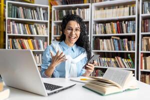 glad ung kvinna i glasögon engagerar animatedly över en smartphone ringa upp, omgiven förbi hyllor fylld med böcker i en bibliotek miljö. foto