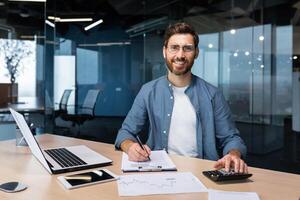porträtt av framgångsrik affärsman Bakom papper arbete, man i skjorta leende och ser på kamera, finansiär chef inuti kontor använder sig av bärbar dator i arbete på arbetsplats. foto