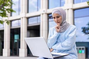 en omtänksam professionell kvinna bär en hijab är sett funderar medan använder sig av henne bärbar dator utanför en modern kontor byggnad. hon är fokuserad, visa upp en blandning av modern professionell liv och kulturell identitet. foto