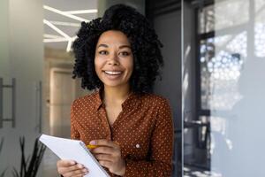 en glad ung kvinna med lockigt hår deltar i ett uppkopplad Träning session, tar anteckningar och leende i en modern kontor miljö. foto