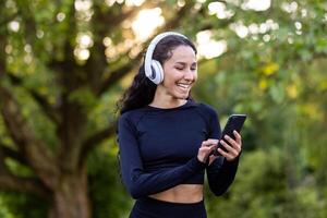 ung skön latin amerikan kvinna promenader i de parkera under aktiva fysisk övningar, kvinna användningar ett Ansökan för lyssnande till audio böcker och musik på telefon, leende sports i hörlurar. foto