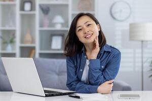 porträtt av ung skön asiatisk kvinna på Hem, kvinna leende och ser på kamera, arbetssätt avlägset i Hem kontor använder sig av bärbar dator medan Sammanträde på tabell i levande rum. foto