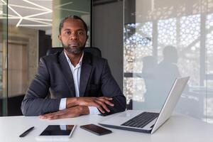 porträtt av allvarlig tänkande och koncentrerad chef, afrikansk amerikan man ser på kamera med vikta händer på skrivbord, chef arbetssätt med bärbar dator inuti kontor, investerare självsäker av seger. foto