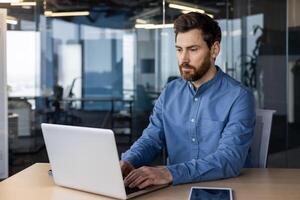 allvarlig professionell manlig i en blå skjorta djupt koncentrerad på arbete använder sig av en bärbar dator i en ljus företags- kontor miljö. foto