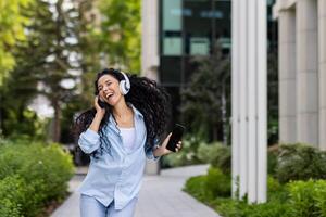 ung glad skön kvinna dans och sång i hörlurar medan gående i de stad, latinamerikan kvinna med lockigt hår användningar ett Ansökan på de telefon, till lyssna till musik uppkopplad. foto