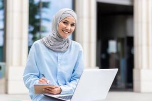 en ung muslim kvinna fokuserade på studerar utanför, tar anteckningar bredvid henne bärbar dator på en universitet campus. foto