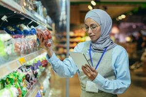 en muslim kvinna i en hijab, en mataffär arbetstagare checkar Produkter på hyllor och i kylskåp, en kvinna med en läsplatta och ett förkläde användningar en läsplatta till recension Produkter foto