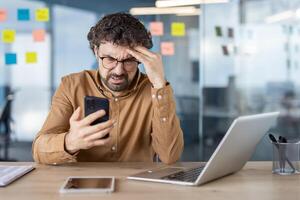 latinamerikan affärsman utseende förvirrad och bedrövad medan använder sig av hans smartphone på en skrivbord med en bärbar dator i en modern kontor miljö. foto