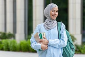 professionell ung arab affärskvinna i hijab leende medan innehav böcker, gående förbi ett kontor byggnad i de stad. foto