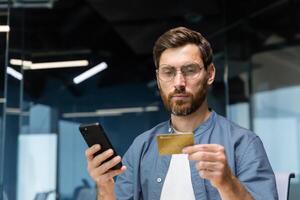 allvarlig tänkande man inuti kontor innehav Bank kreditera kort och smartphone, affärsman framställning pengar överföra, bokning och inköp i uppkopplad Lagra, framgångsrik entreprenör. foto