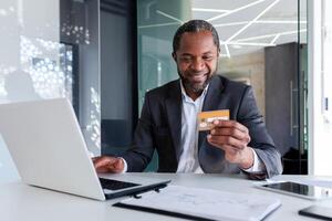 afrikansk amerikan leende manlig affärsman arbetssätt i kontor på bärbar dator och innehav och använder sig av kreditera kort. foto
