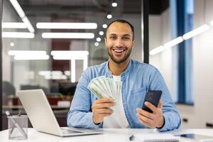 porträtt av en Lycklig ung latinamerikan man Sammanträde i de kontor på de tabell, innehav kontanter pengar och en telefon, leende på de kamera. foto