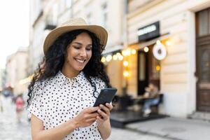 en skön ung kvinna promenader genom de kväll stad i en hatt, en leende latin amerikan kvinna innehar en smartphone i henne händer. en turist med lockigt hår typer en meddelande och bläddrar uppkopplad sidor på de telefon. foto