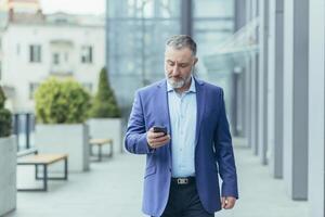 gråhårig senior stilig man affärsman i kostym gående nära kontor Centrum på gata, innehav mobil telefon, skriver meddelande, kontroll post. foto