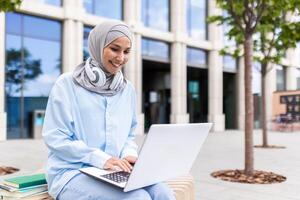 ung muslim kvinna bär en hijab arbetssätt på en bärbar dator medan Sammanträde på en bänk utanför högskola byggnader. foto