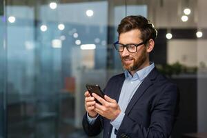 framgångsrik finansiär investerare Arbetar inuti kontor på arbete, affärsman i företag kostym användningar telefon nära fönster, man ler och läser Bra Nyheter uppkopplad från smartphone. foto