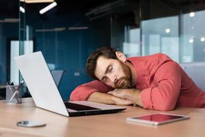 trött affärsman i tillfällig röd skjorta sover på arbetsplats, man på skrivbord föll sovande under arbetssätt timmar inuti kontor med bärbar dator. foto