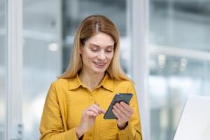 mogna erfaren företag kvinna inuti kontor på arbetsplats, anställd Sammanträde på skrivbord leende innehav telefon i hand, chef använder sig av Ansökan på smartphone, bläddring internet sidor. foto