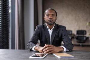 en självsäker afrikansk amerikan affärsman sitter på en skrivbord i en väl upplyst modern kontor, ser direkt på de kamera, utsöndrar professionalism och fokus. foto