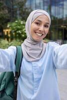 glad kvinna bär en hijab fångar en vertikal selfie, eventuellt under en ringa upp, med ett urban bakgrund. hon utstrålar en personligt och engagerande närvaro. foto