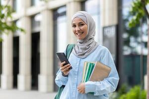 leende arab kvinna i hijab innehav en smartphone och anteckningsbok utanför ett kontor byggnad, utsöndrar förtroende och professionalism. foto