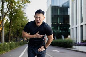 asiatisk ung manlig sportsman står böjd över på de stad gata och innehar hans hand till hans bröst, känt svår smärta och spasm i hans hjärta. foto