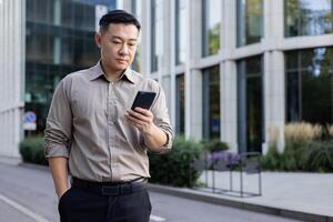 allvarlig ung asiatisk manlig advokat stående nära kontor byggnad och använder sig av mobil telefon. foto