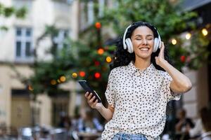 ung skön latinamerikan kvinna lyssnar till musik sjunger längs och danser medan gående i kväll stad, kvinna med lockigt hår användningar hörlurar och app på telefon. foto