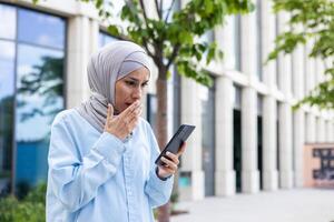 ung företag kvinna i hijab gående i de stad, arab kvinna mottagen uppkopplad underrättelse med dålig Nyheter, innehav telefon i händer läsning upprörd och deprimerad ledsen. foto