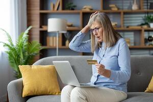 en mogna kvinna ser överraskad och angelägen medan innehav en kreditera kort och använder sig av en bärbar dator på henne soffa på Hem. foto