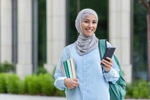 ung muslim kvinna i en hijab innehav böcker och en smartphone, leende utomhus nära ett kontor byggnad, symboliserar bemyndigande. foto