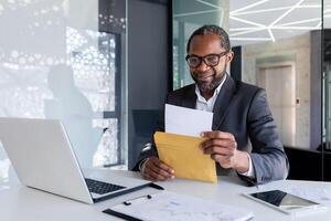 senior nöjd finansiär mottagen en brev post meddelande i ett kuvert, affärsman läser och ler, chef på skrivbord inuti kontor användningar bärbar dator. foto