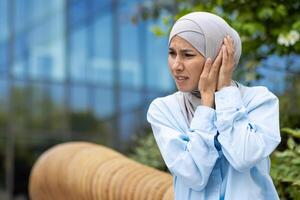 medelålders kvinna i en ljus blå hijab innehav henne öra i smärta utanför, med en bekymrad uttryck, eventuellt lidande från otit eller öra inflammation. foto