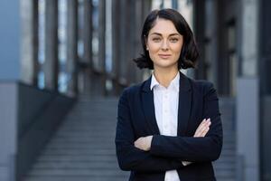 en professionell affärskvinna står självsäkert utanför en modern kontor byggnad, vapen korsade, bär en företag kostym med en positiv uttryck. foto