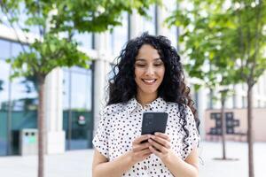 en ung skön kvinna promenader genom de stad med en telefon i henne händer, ler belåtet, användningar en smartphone Ansökan, bläddrar internet sidor, typer en text meddelande, social nätverk. foto