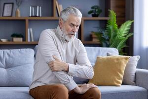 ett äldre man upplever obehag innehar hans axel medan sittande på en soffa, illustrerar hälsa frågor i senior levande. foto