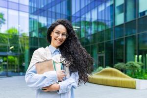 porträtt av ung skön latin amerikan kvinna studerande, kvinna med böcker och bärbara datorer leende och ser på kamera, kvinna lärare utanför universitet campus kontor byggnad. foto