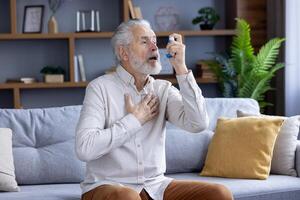 äldre caucasian man upplever ett astma ge sig på, använder sig av en blå inhalator medan Sammanträde på en soffa i en modern levande rum. foto