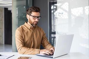 allvarlig koncentrerad tänkande affärsman arbetssätt inuti kontor med bärbar dator, mogna upplyst man i skjorta och glasögon på arbetsplats skriver på tangentbord, programmerare utvecklaren. foto