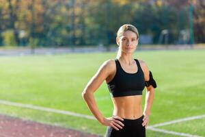 porträtt av mogna vuxen kvinna idrottare i stadion, kvinna idrottare innehav händer på höfter leende och ser på kamera, joggning kvinna i träningsoverall under aktiva övning och kondition klass. foto